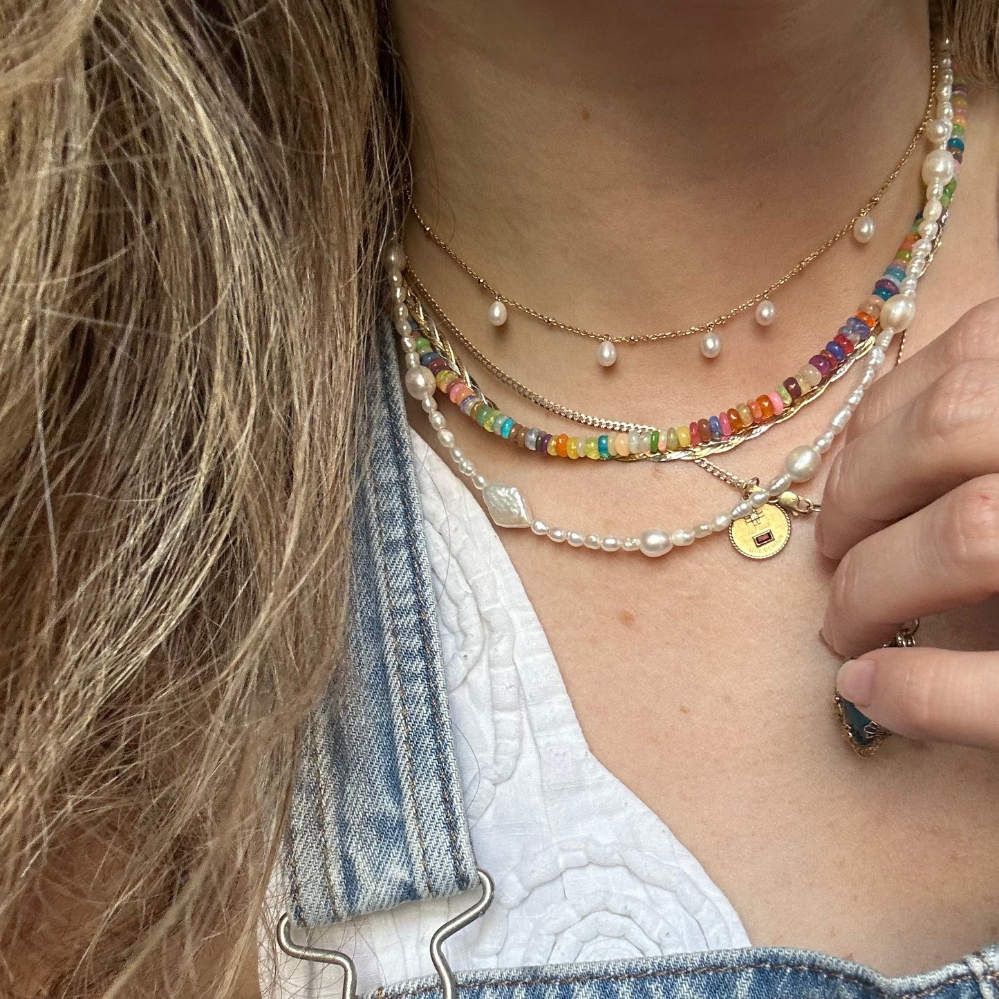 A close-up of a person wearing four layered necklaces, including one with the Solid Gold Coin Charm. The person is dressed in a white top and denim.