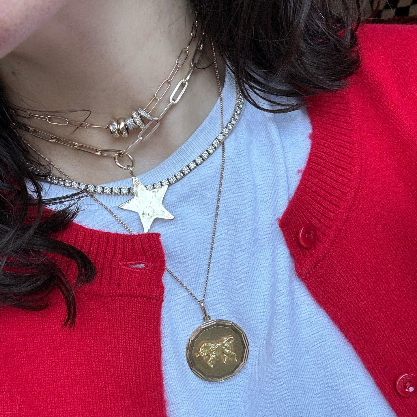 A close-up shows a person in a white shirt and red sweater wearing layered gold Luiny x Local Eclectic Charms necklaces, featuring an exclusive pendant and round coin. Their dark hair is partially visible, highlighting the unique jewelry style.