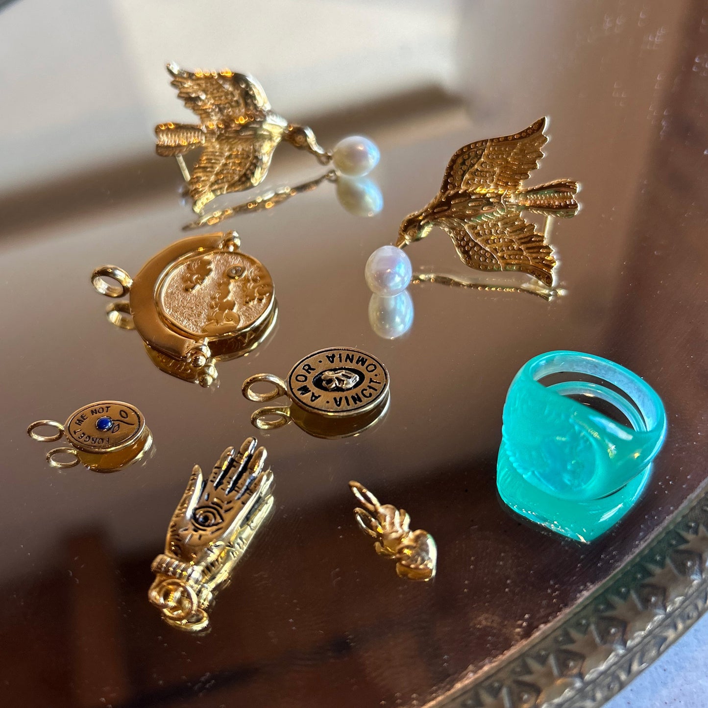Charms by Hunt of Hounds—gold vermeil birds, moon, hand, medallion, and text charms—are displayed on a reflective surface with pearls on two bird charms, alongside a chunky turquoise ring.