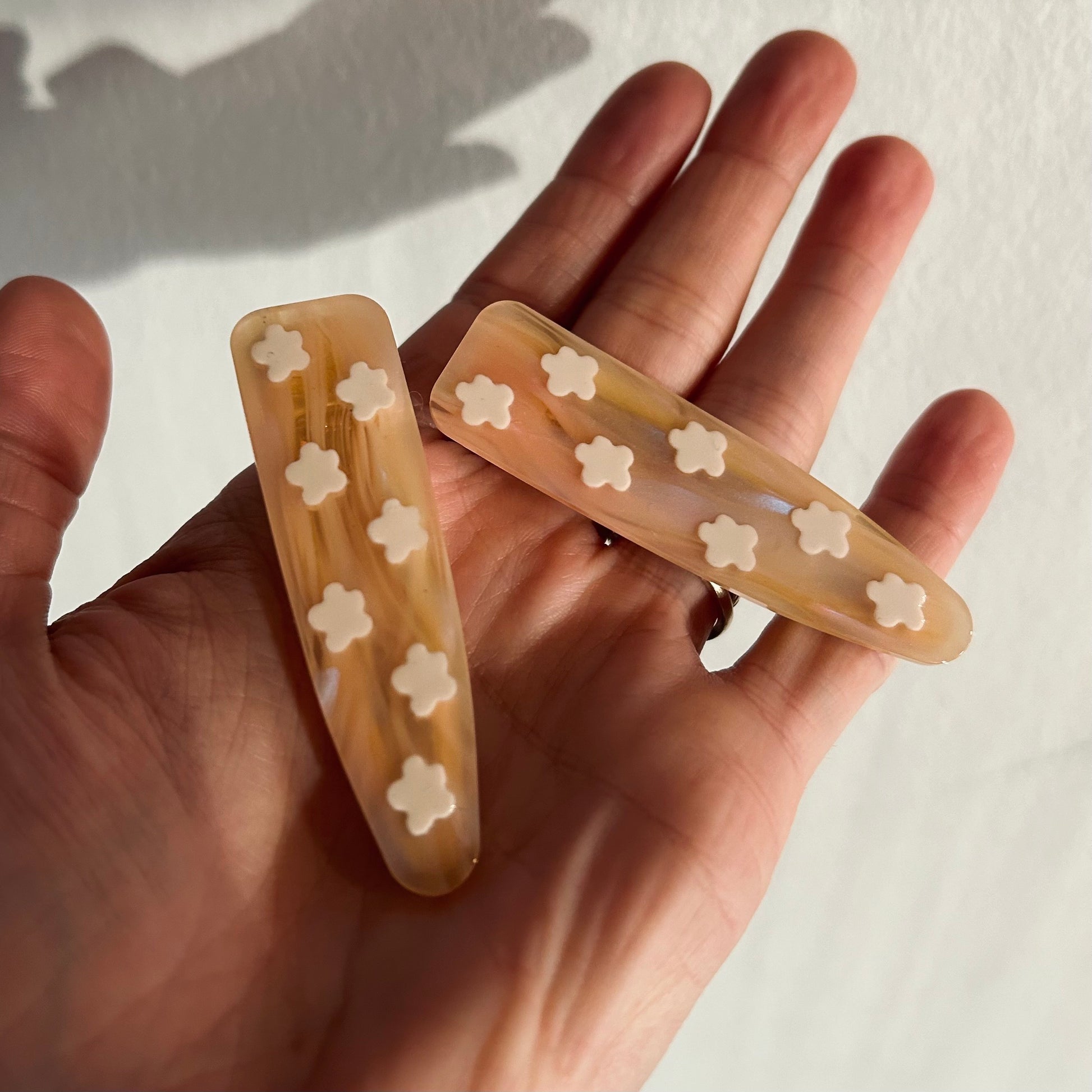 A hand holds two beige resin hair clips from the Flower Barrette Set, each adorned with small white flower accents against a sunlit white background—charming floral accessories for any occasion.