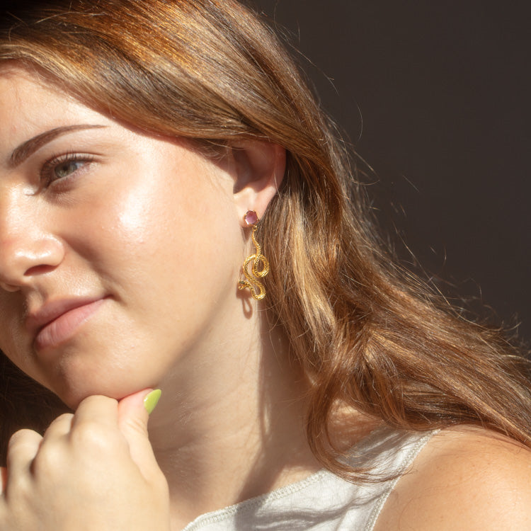 A young woman with long brown hair wears the Awaken Amethyst Snake Earrings—18k gold plated drop earrings featuring a small pink stone. She gazes thoughtfully to the side, chin resting on her hand in soft sunlight.