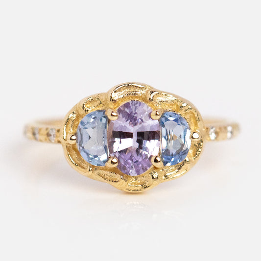 The Periwinkle Ring features an oval-cut light purple gemstone at the center, accented by two light blue ovals, all set in a detailed 14k yellow gold setting with a sleek band.