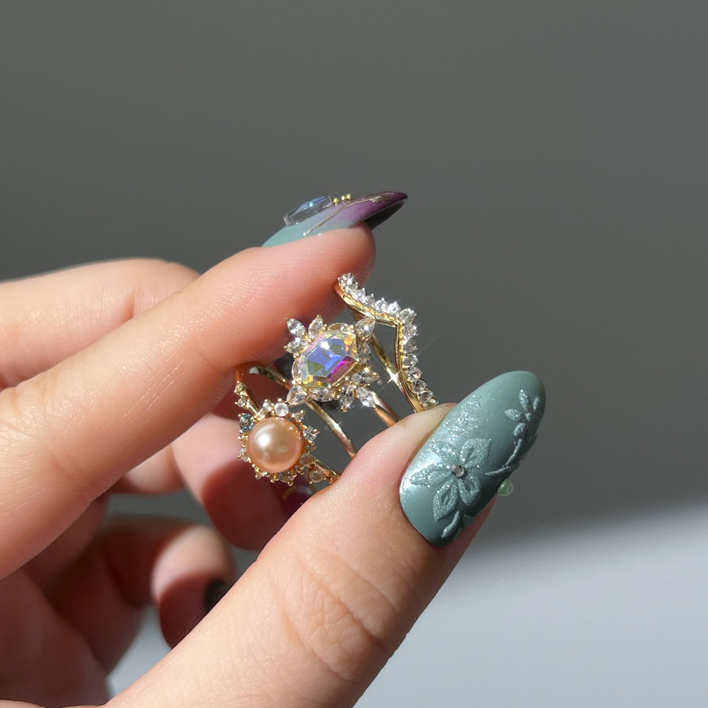 A hand with teal floral nail art holds the 14k Pastel Pink Pearl Statement Ring, featuring crystals and gemstones, illuminated by sunlight against a neutral background.