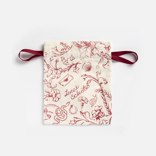 The Cotton Jewelry Pouch is small and cream-colored, has a grosgrain ribbon closure, detailed red prints of animals, plants, shells, and features the words Loca Collecto.