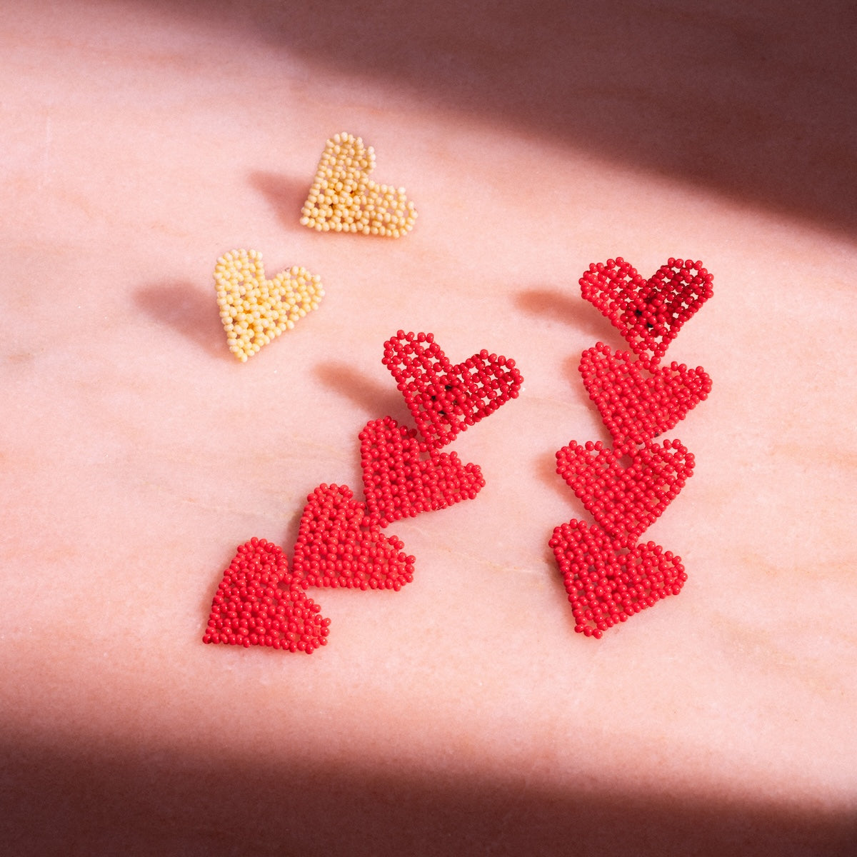 Eight beaded heart-shaped pins, similar to the Dae Earring, rest on pink marble: six red hearts aligned in two rows and two yellow hearts—like bright yellow Dae Earrings—side by side above them.