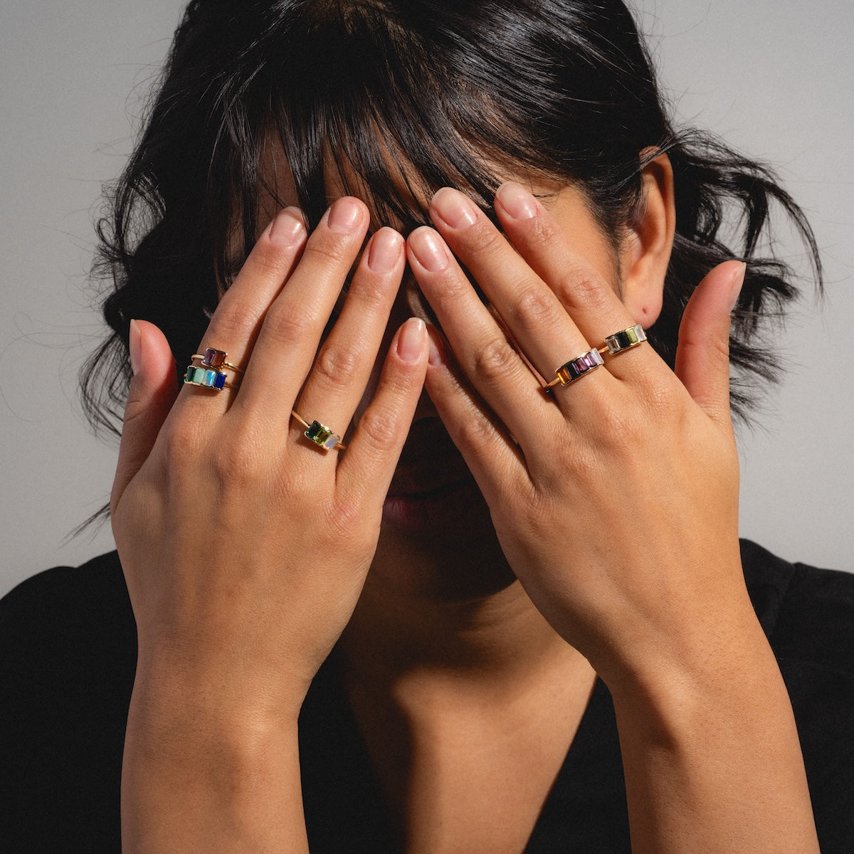 A person with dark hair and a black top covers their face with both hands, displaying several colorful rings, including the Customizable Acrostic Baguette Gemstone Ring, against a plain background.