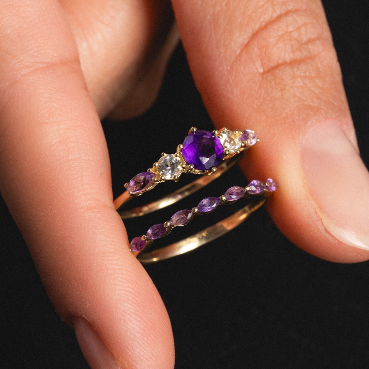 A hand displays the Solid Gold 2025 February Capsule Classic Amethyst Statement Ring—featuring a gold double band with purple and clear gemstones—set against a black background.