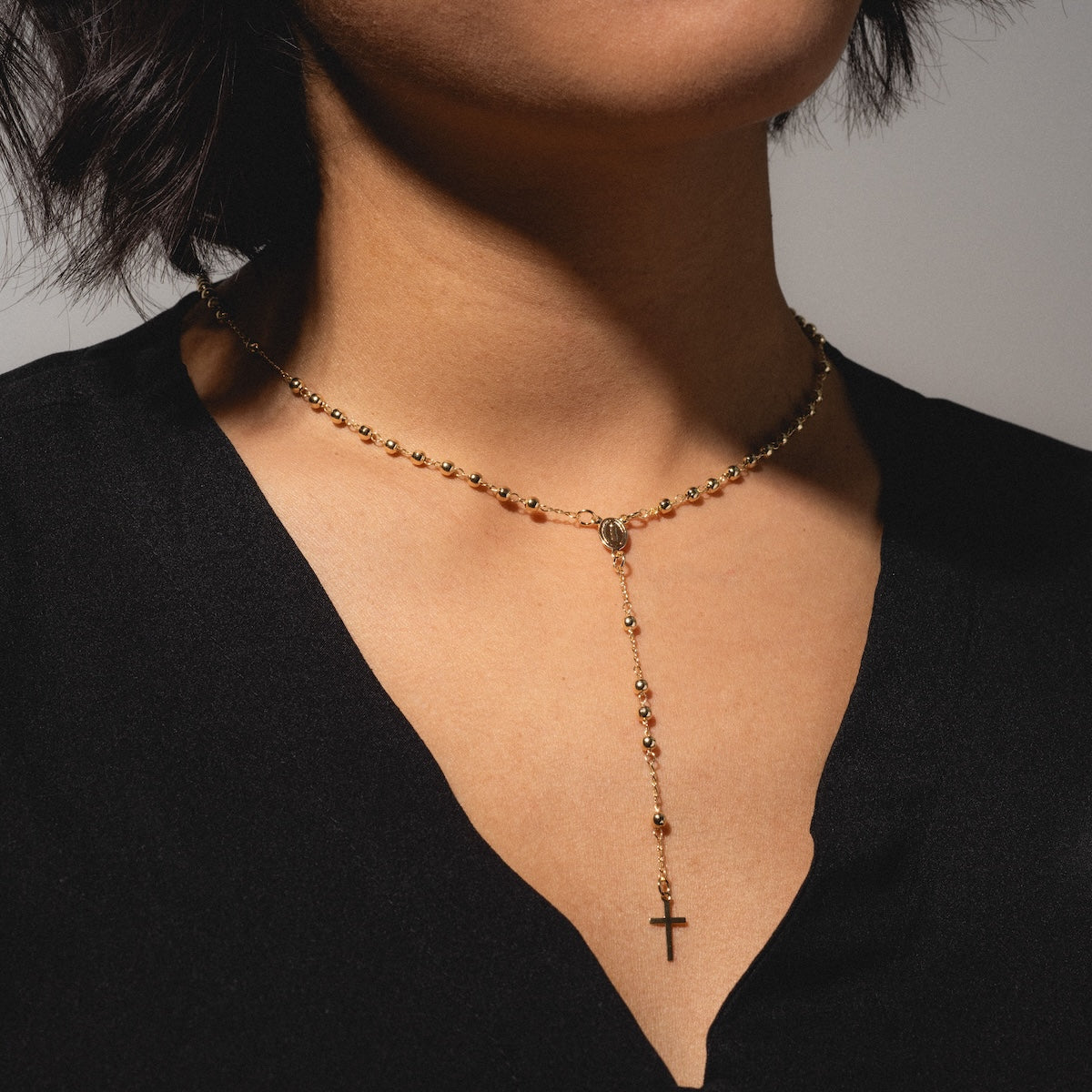 A person wearing a black V-neck top and a Gold Rosary Necklace—a 14k gold plated beaded chain ending in a gold cross pendant. The face is partially out of frame.