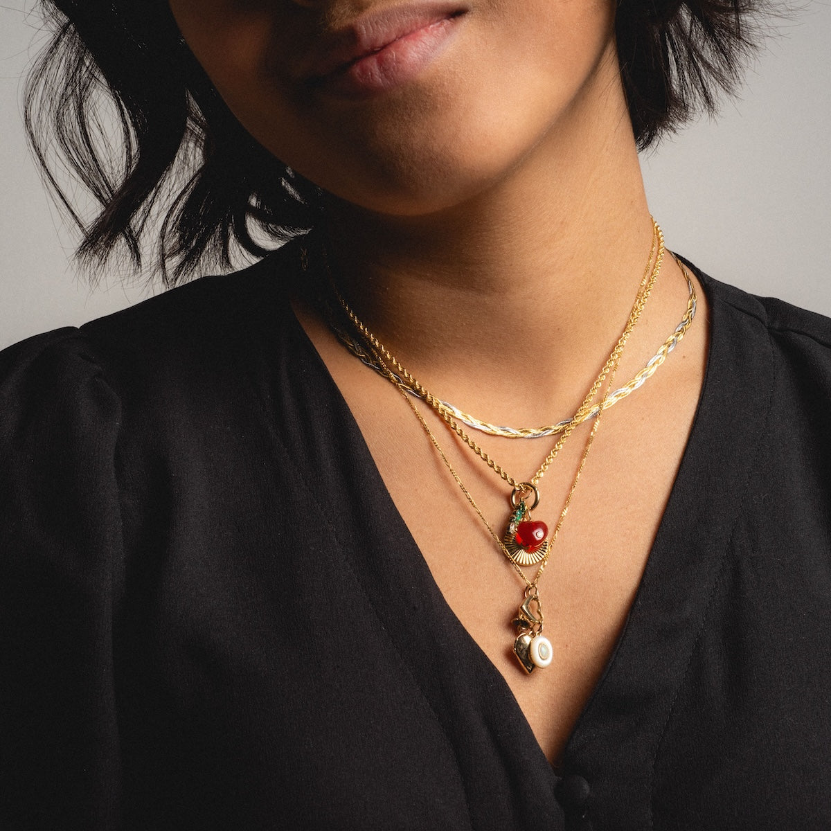 A woman, shown from the shoulders up with a subtle smile, wears a black v-neck top and layered gold necklaces, including the Solid Gold Mixed Metal Braided Necklace. Her face is partially visible.
