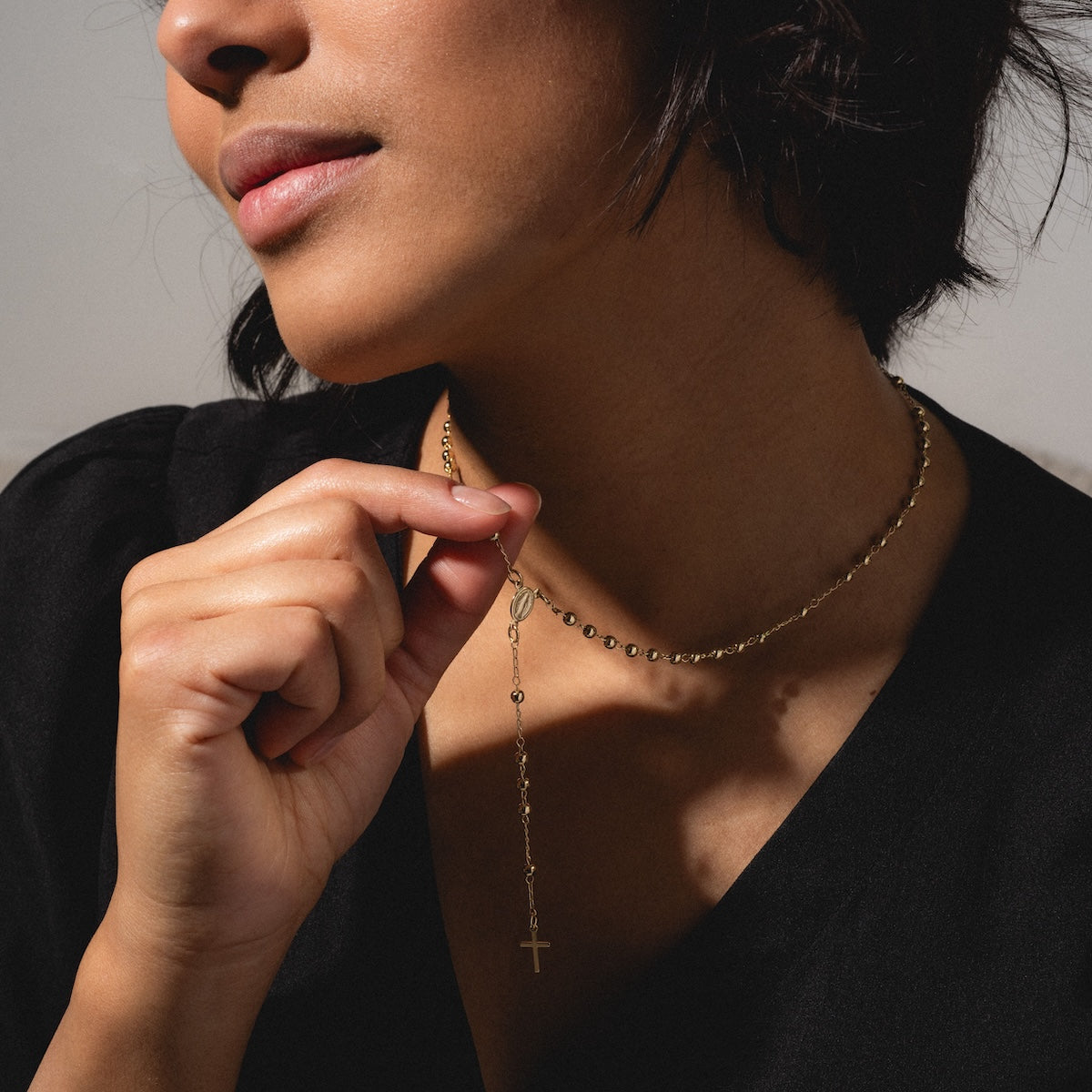 A woman in a black top holds the Gold Rosary Necklace—an elegant 14k gold plated piece with a small cross pendant—close to her neck. Her face is partially visible, softly lit to highlight this graceful faith jewelry.