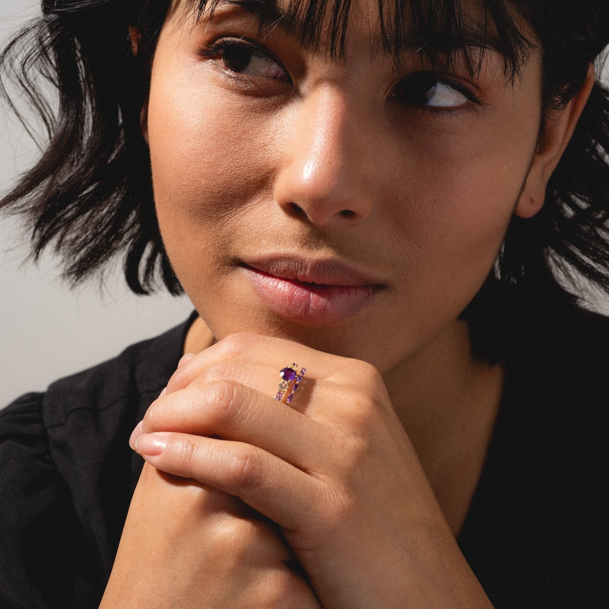A woman with short dark hair, dressed in a black top, rests her chin on her hand while wearing the Solid Gold 2025 February Capsule Classic Amethyst Statement Ring, adorned with small purple stones, and gazes thoughtfully to the side.