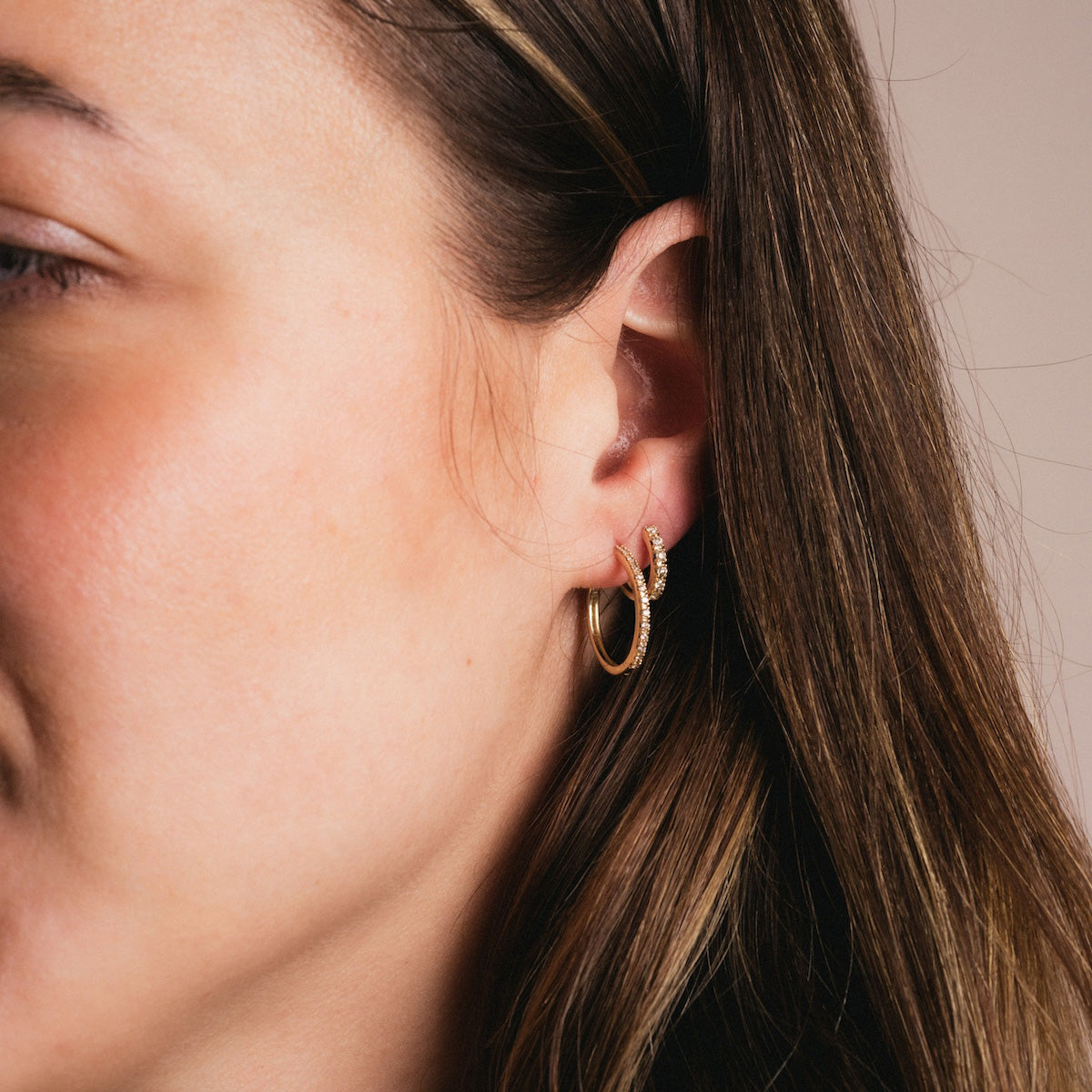 Close-up of a womans ear with long brown hair, wearing two Solid Gold Diamond Hoops and a small gold ear cuff on the upper ear.