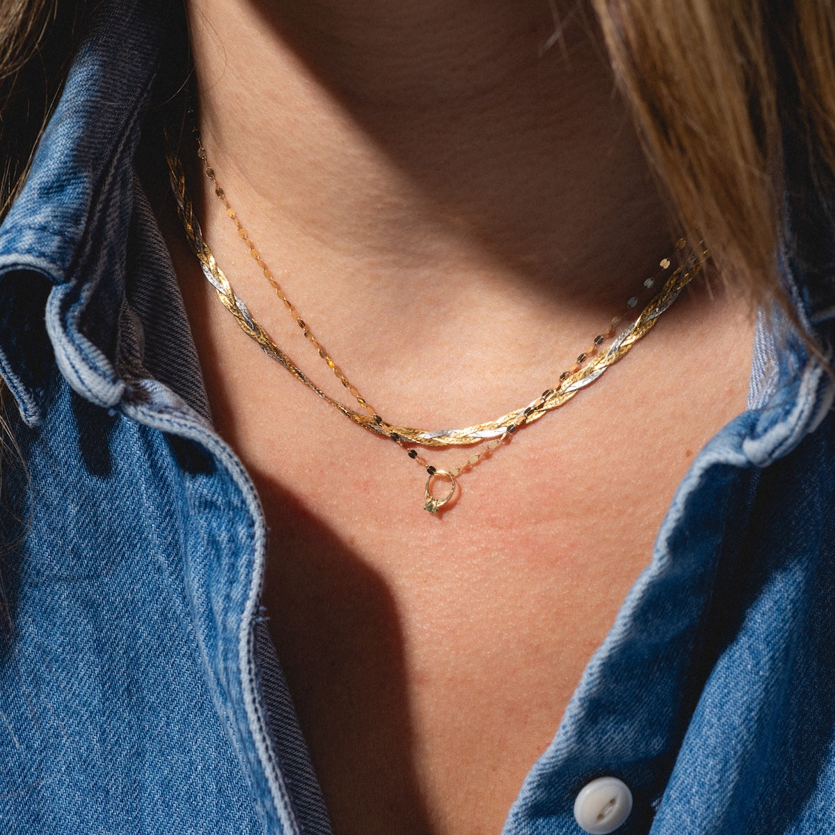 A close-up of a person wearing a blue denim shirt and two layered gold necklaces, featuring the 14k Littlest Birthstone Ring Charm. The open collar lets sunlight highlight the jewelry and skin.