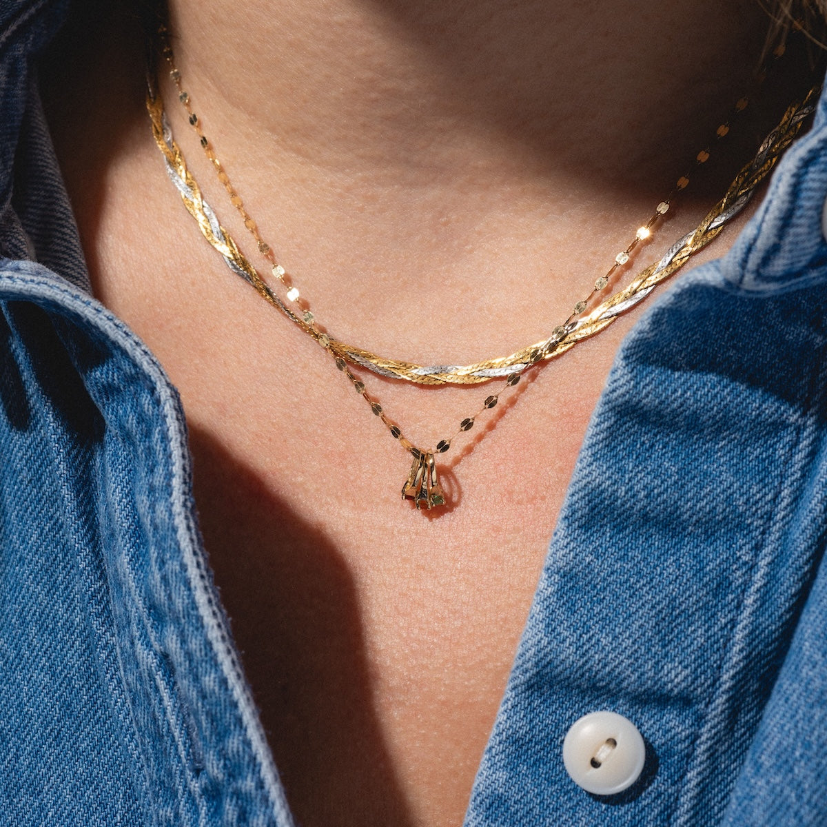 A close-up of a person in a blue denim shirt, partially unbuttoned to reveal two layered gold necklaces. One features the 14k Littlest Birthstone Ring Charm by Charlie and Marcelle on their neck.
