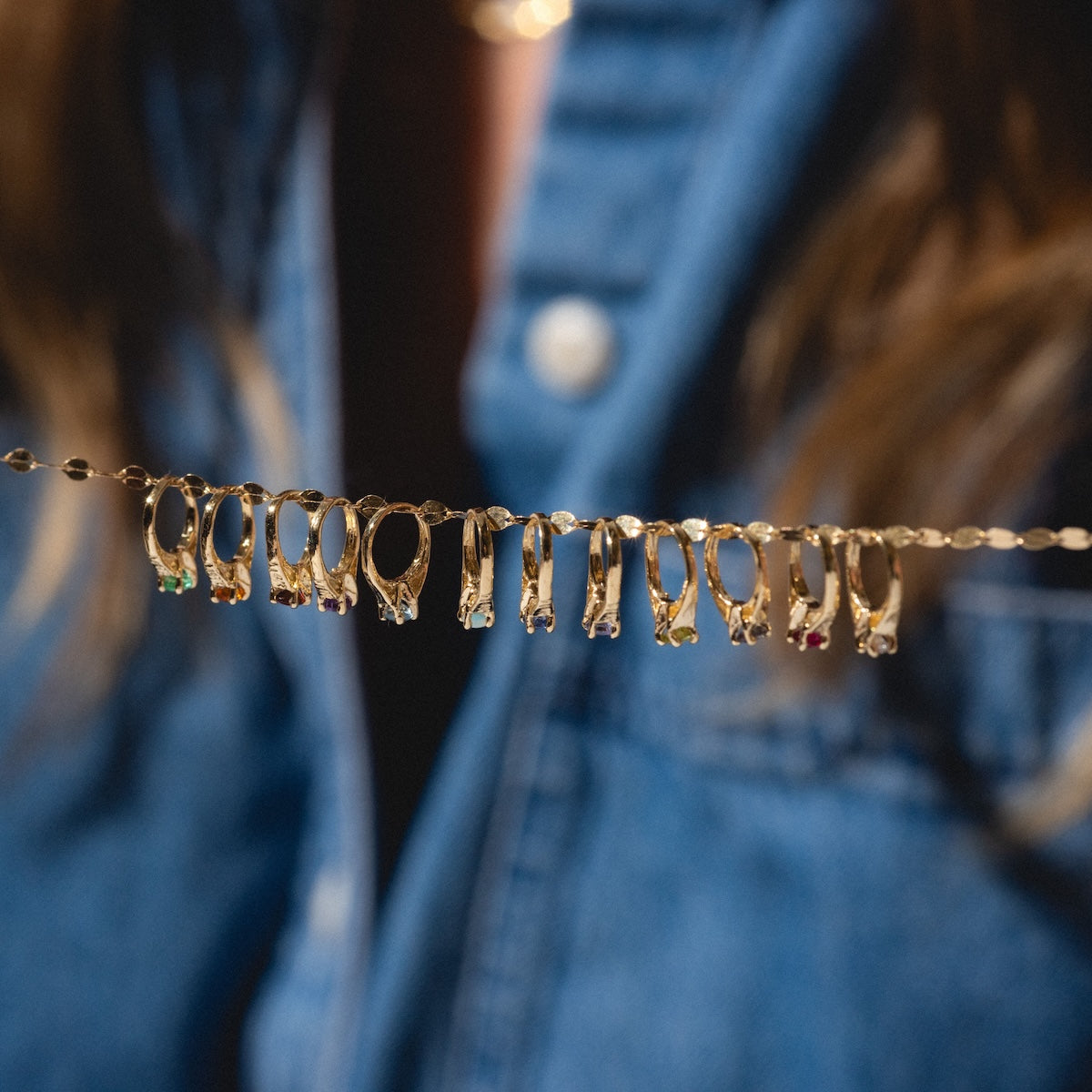 A close-up of a 14k Littlest Birthstone Ring Charm featuring colorful stones, attached to a gold chain necklace. In the background, someone wears a blue denim shirt.