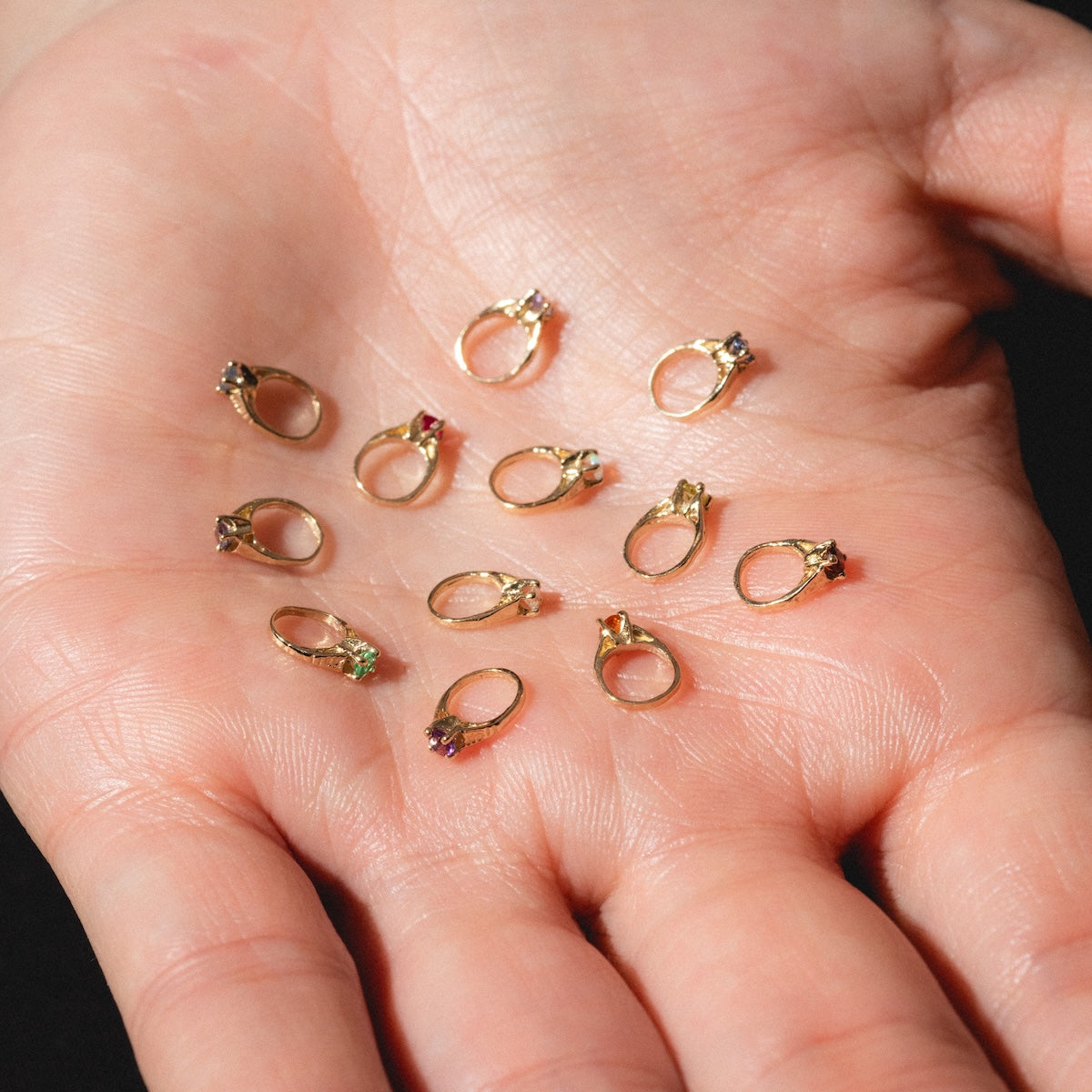 A hand displays several small gold rings, including the 14k Littlest Birthstone Ring Charm, each featuring different colored gemstones by Charlie and Marcelle, resting on the palm against a dark background.
