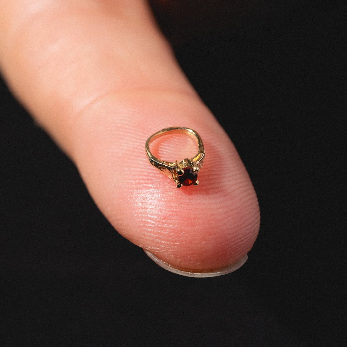 The 14k Littlest Birthstone Ring Charm by Charlie and Marcelle, a tiny gold ring with a red gemstone, is balanced on the tip of a finger against a black background.