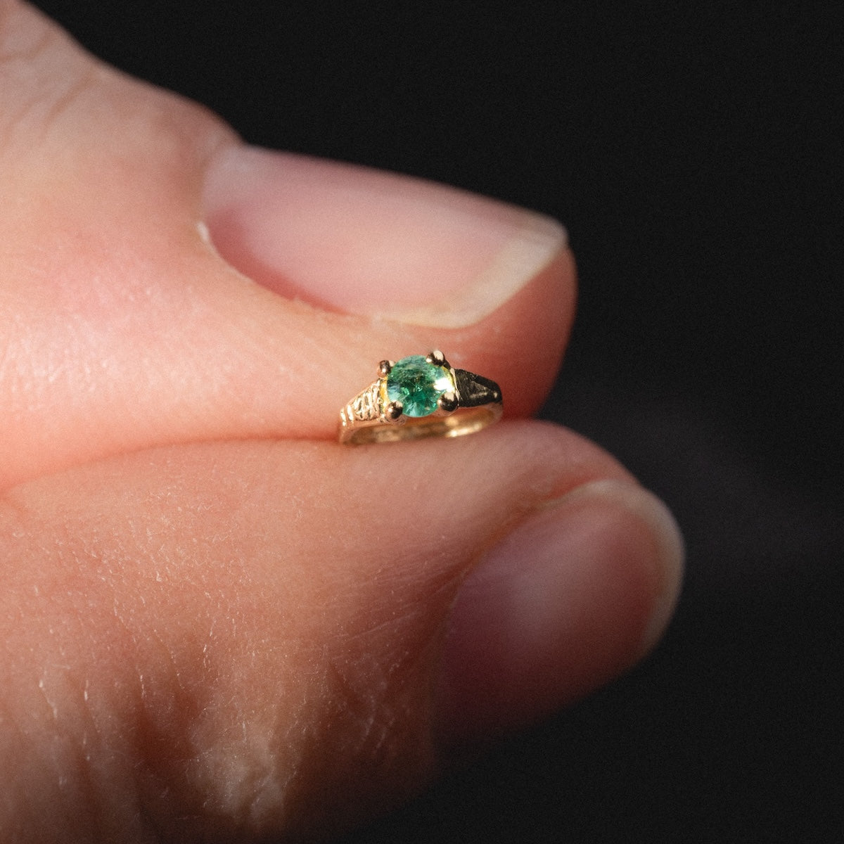 A close-up of fingers holding a 14k Littlest Birthstone Ring Charm with a green gemstone at its center, set against a dark background.