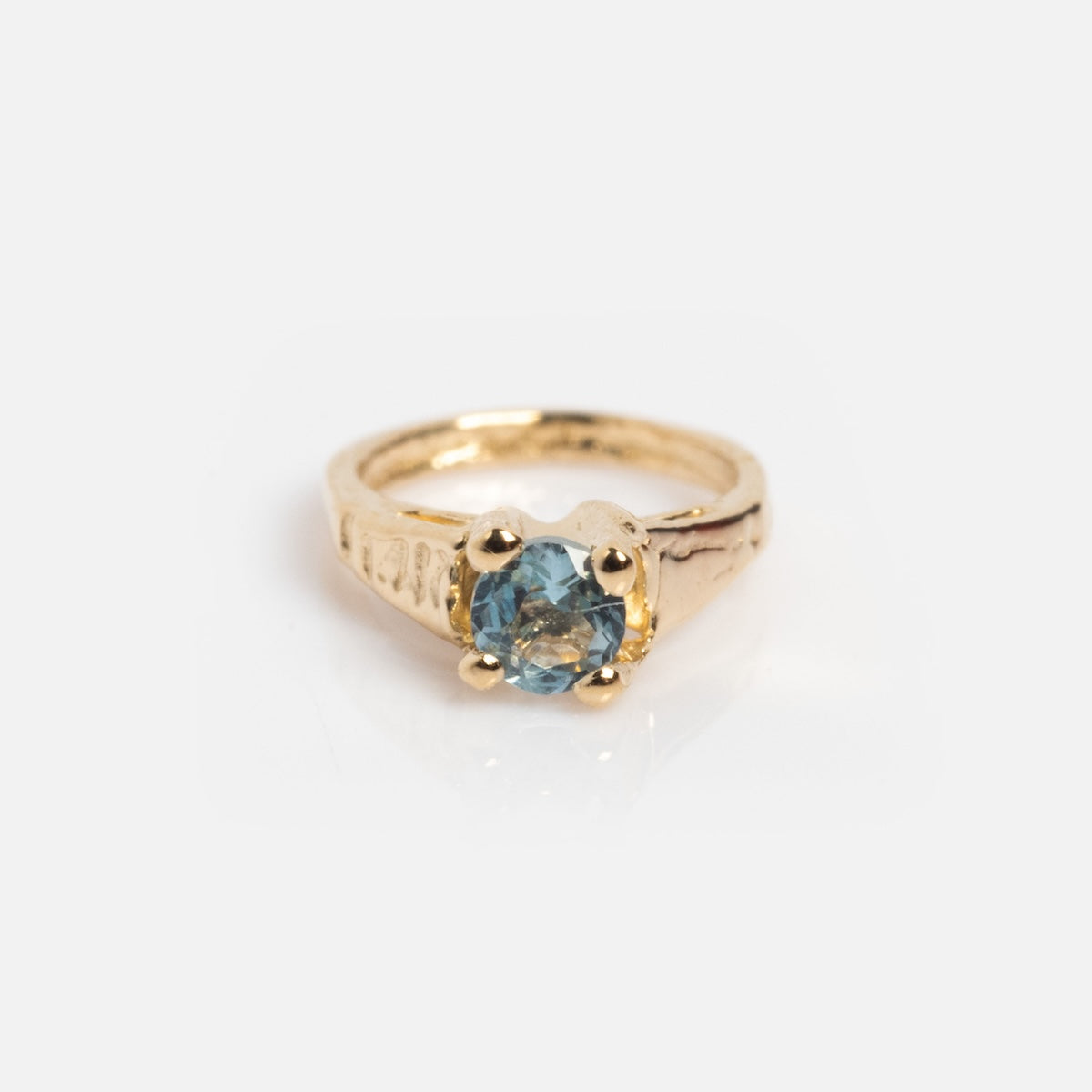The 14k Littlest Birthstone Ring Charm by Charlie and Marcelle features a gold textured band with a round light blue gemstone in a prong setting, elegantly showcased on a white background.