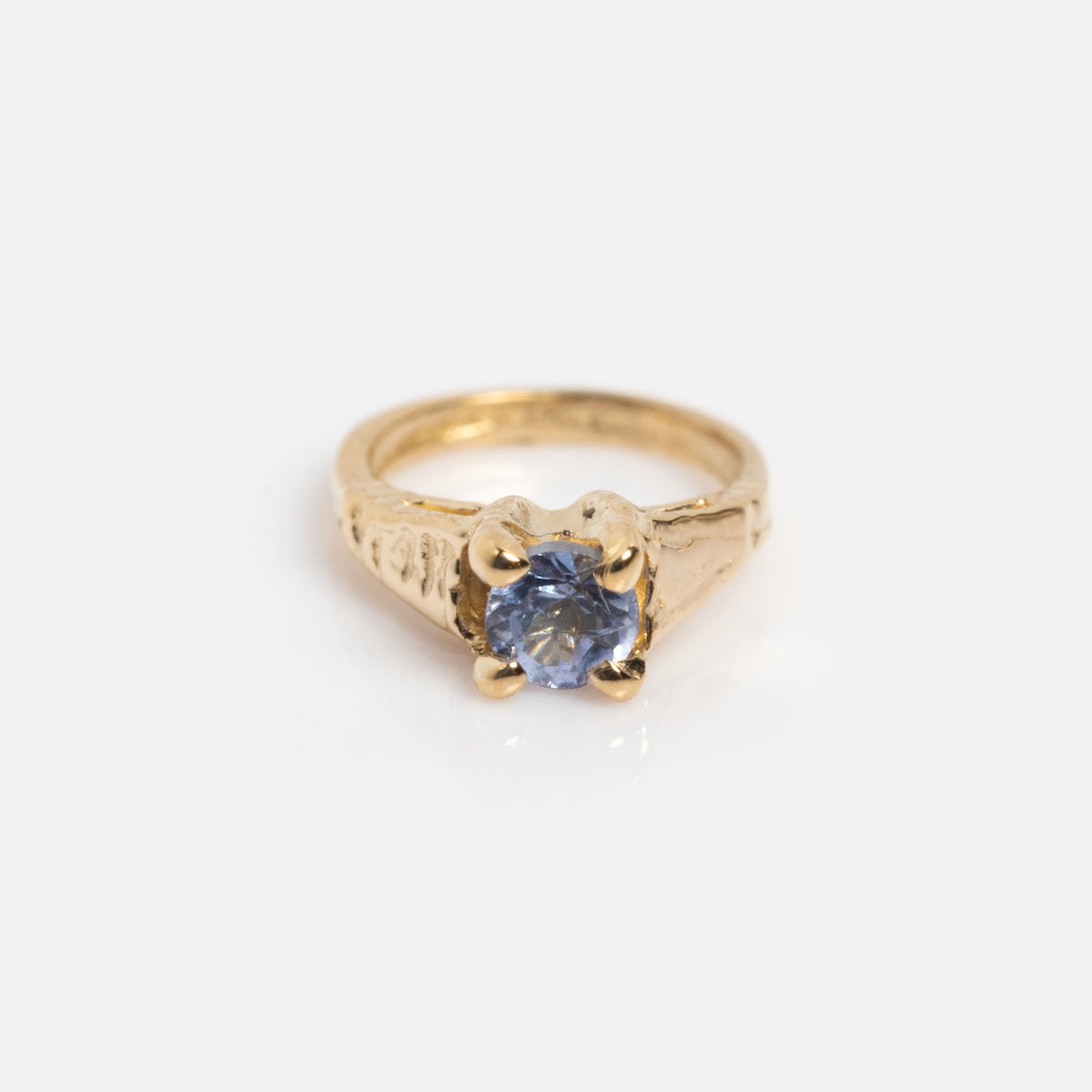 A gold ring with engraved details features a single round blue gemstone in a four-prong setting, reminiscent of the 14k Littlest Birthstone Ring Charm by Charlie and Marcelle, shown on a white background.