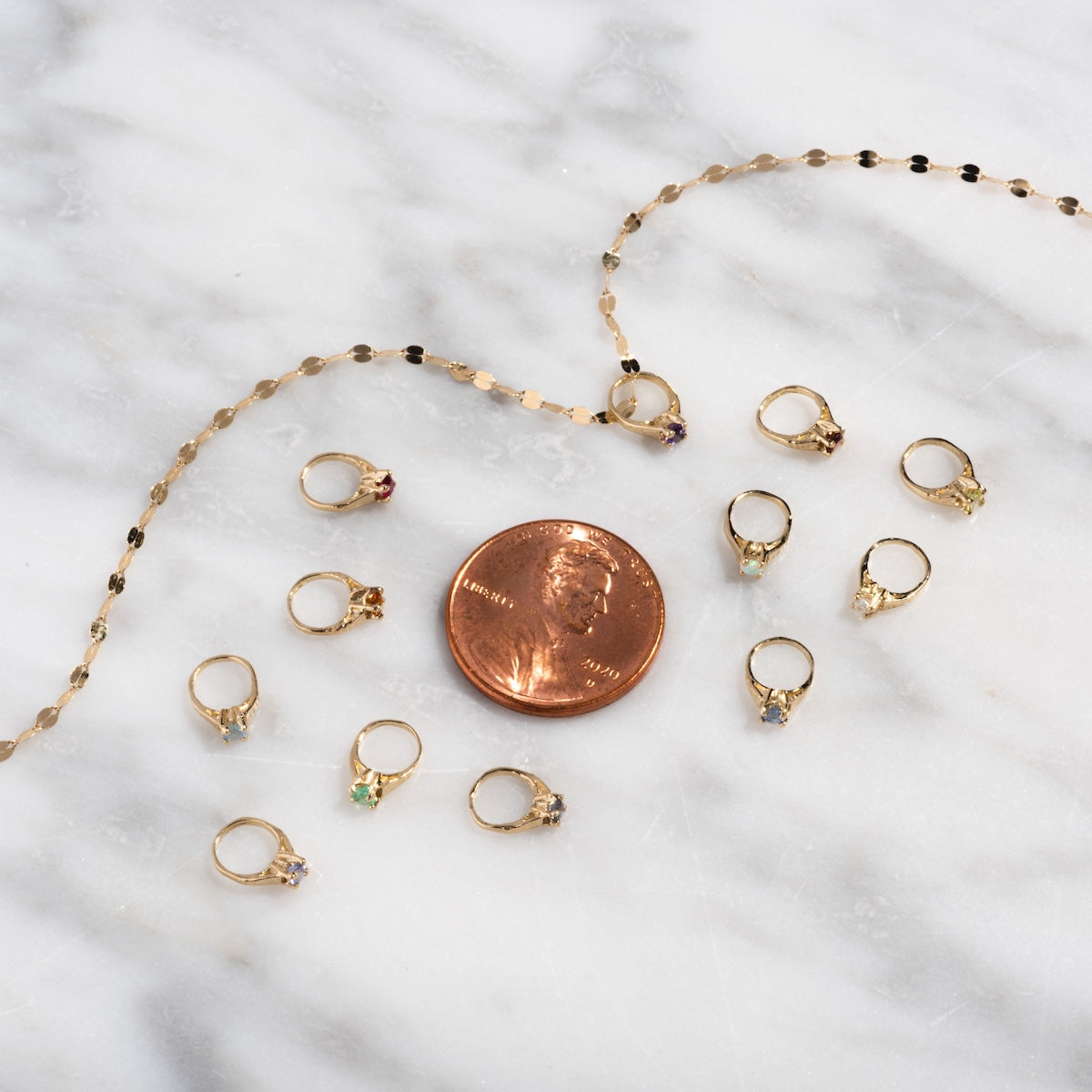 A penny sits beside a solid gold necklace charm, a gold chain, and ten colorful 14k Littlest Birthstone Ring Charms by Charlie and Marcelle on white marble—the penny offers scale for these elegant jewelry pieces.