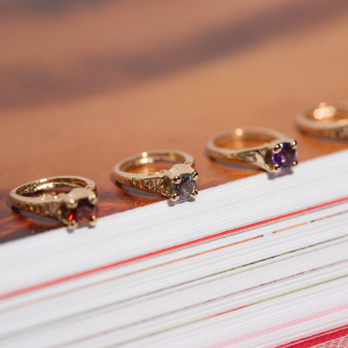 Four gold rings with various gemstones, including the 14k Littlest Birthstone Ring Charm, are lined up on the edge of a thick closed book with visible pages, basking in sunlight.