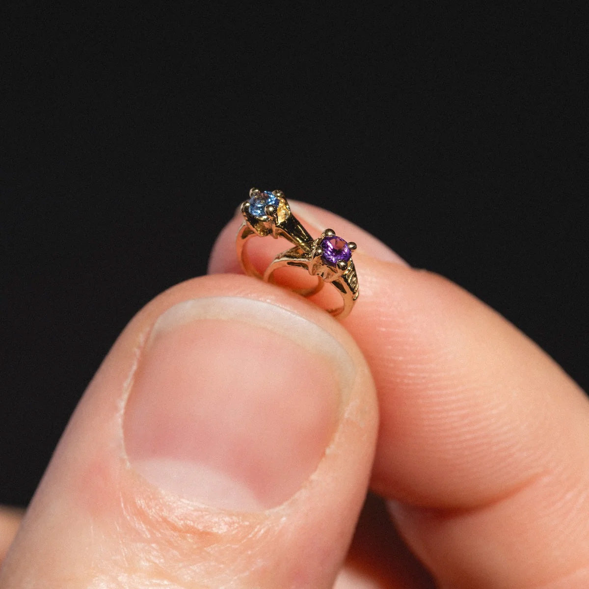 A close-up of a hand holding the 14k Littlest Birthstone Ring Charm, featuring two stones—one blue and one purple—each set in an ornate setting, shown against a black background.