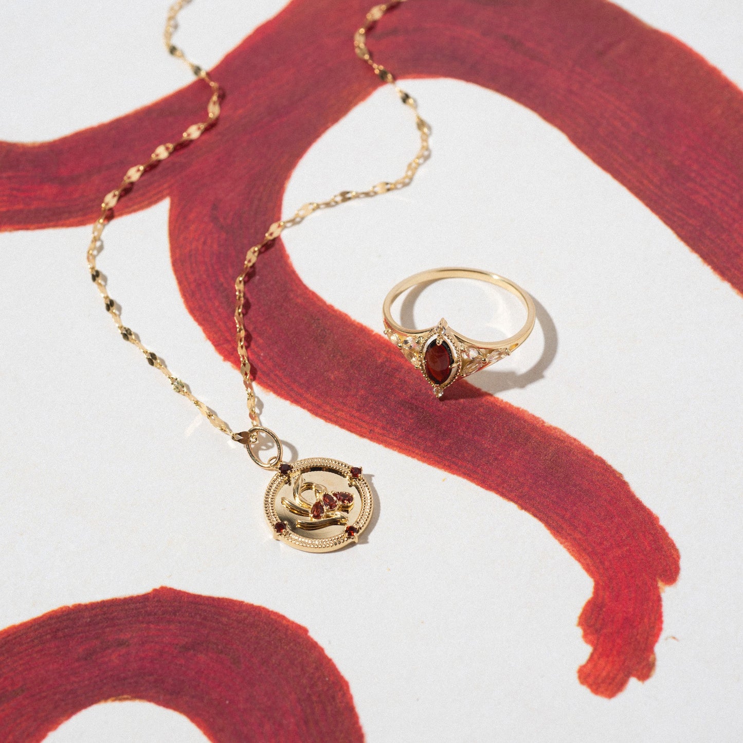 The Solid Gold 2026 January Garnet Capsule Set, featuring a gold necklace with a circular pendant and a vintage-inspired garnet ring, is displayed on white paper with bold red brushstrokes. Free gift included.