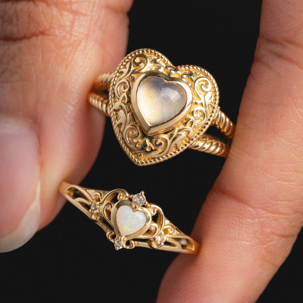 A hand holds two Solid Gold Openwork Filigree Opal Heart Rings, each with ornate heart-shaped filigree designs and Australian opal center stones, shown against a black background.