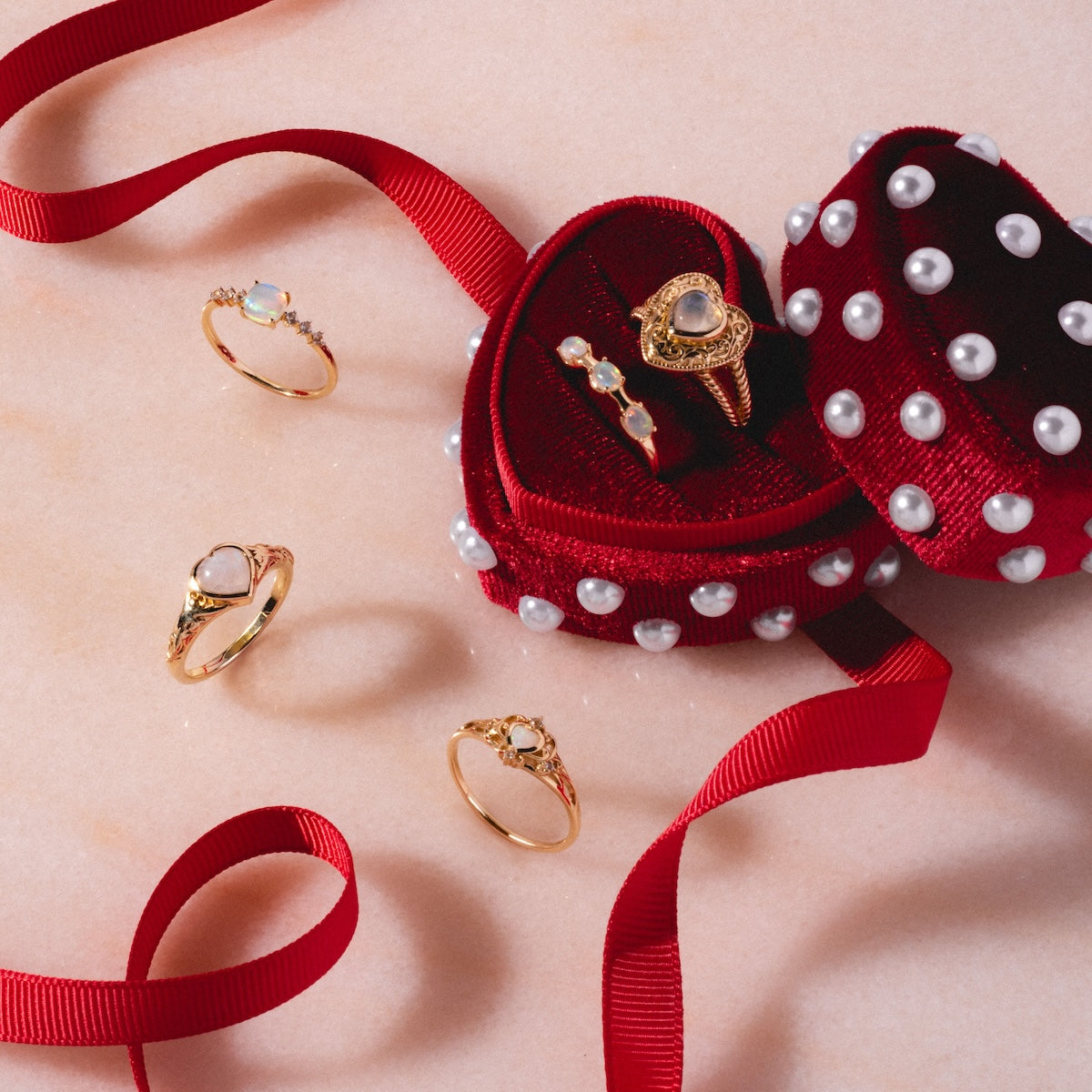 A red, heart-shaped velvet jewelry box with pearls holds a Solid Gold Openwork Filigree Opal Heart Ring. More gold rings with gemstones, including an Australian opal and white topaz accents, are displayed on pink marble with a red ribbon.