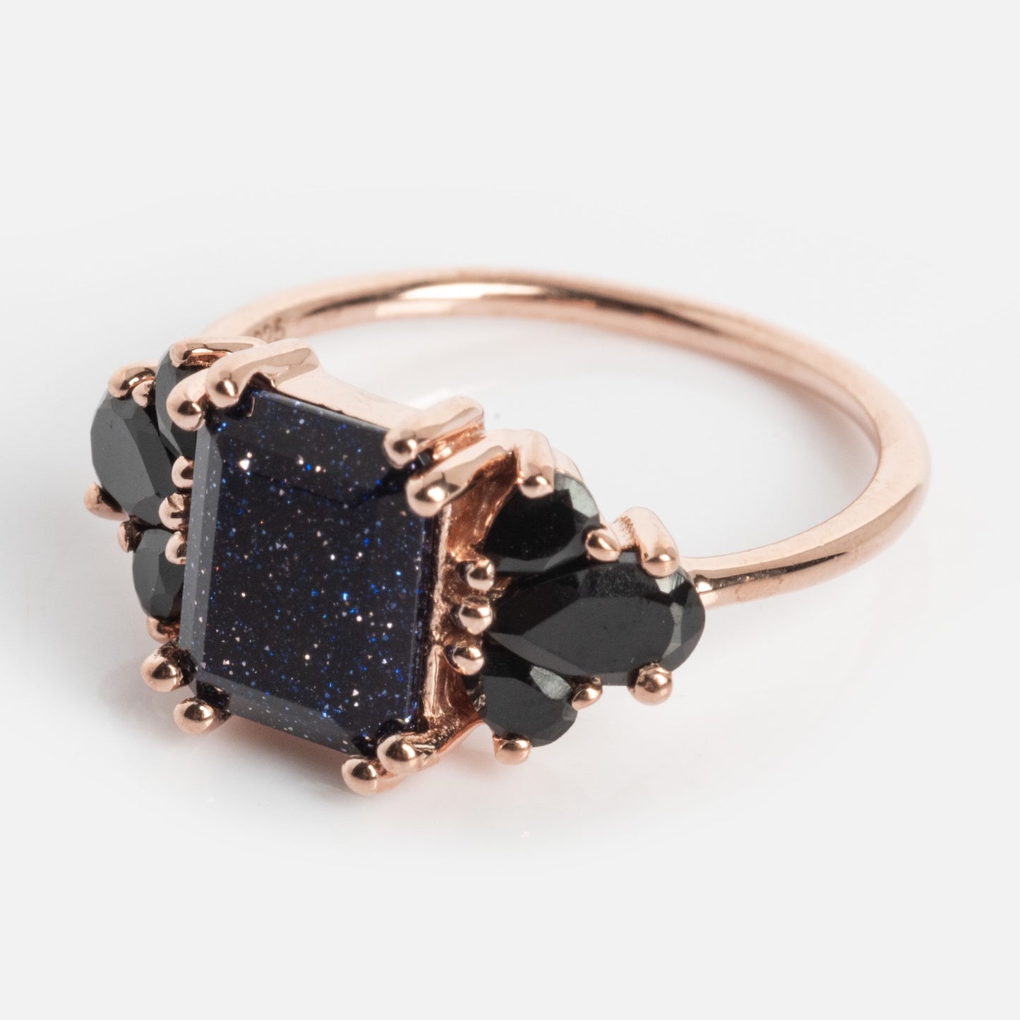 The Gemstone Empress Ring features a rose gold band with a large rectangular dark blue stone flecked with glitter, accented by black teardrop stones on each side, displayed against a plain light background.