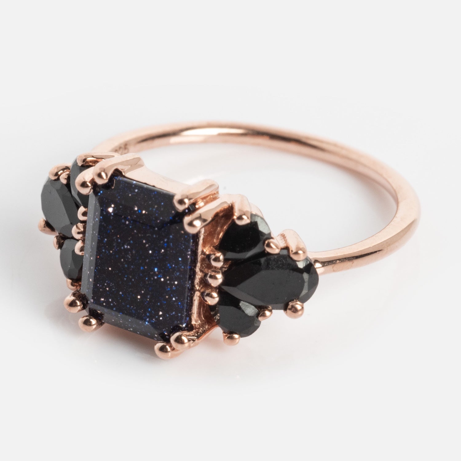 The Gemstone Empress Ring features a rose gold band with a large rectangular dark blue stone flecked with glitter, accented by black teardrop stones on each side, displayed against a plain light background.