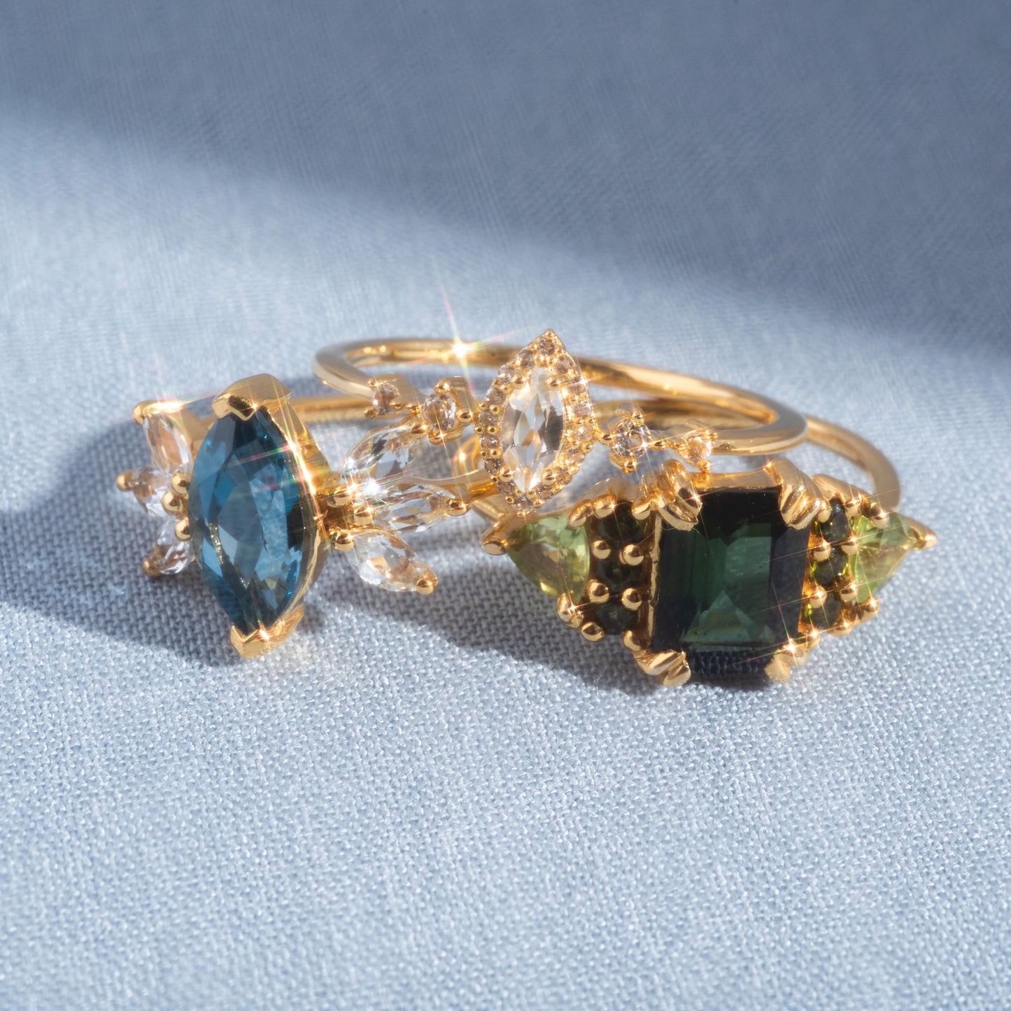 The Green with Envy Topaz and Peridot Regal Ring, a 14k yellow gold vermeil piece with green topaz and clear accents, is displayed alongside a marquise-cut blue stone ring on light blue fabric in bright sunlight.