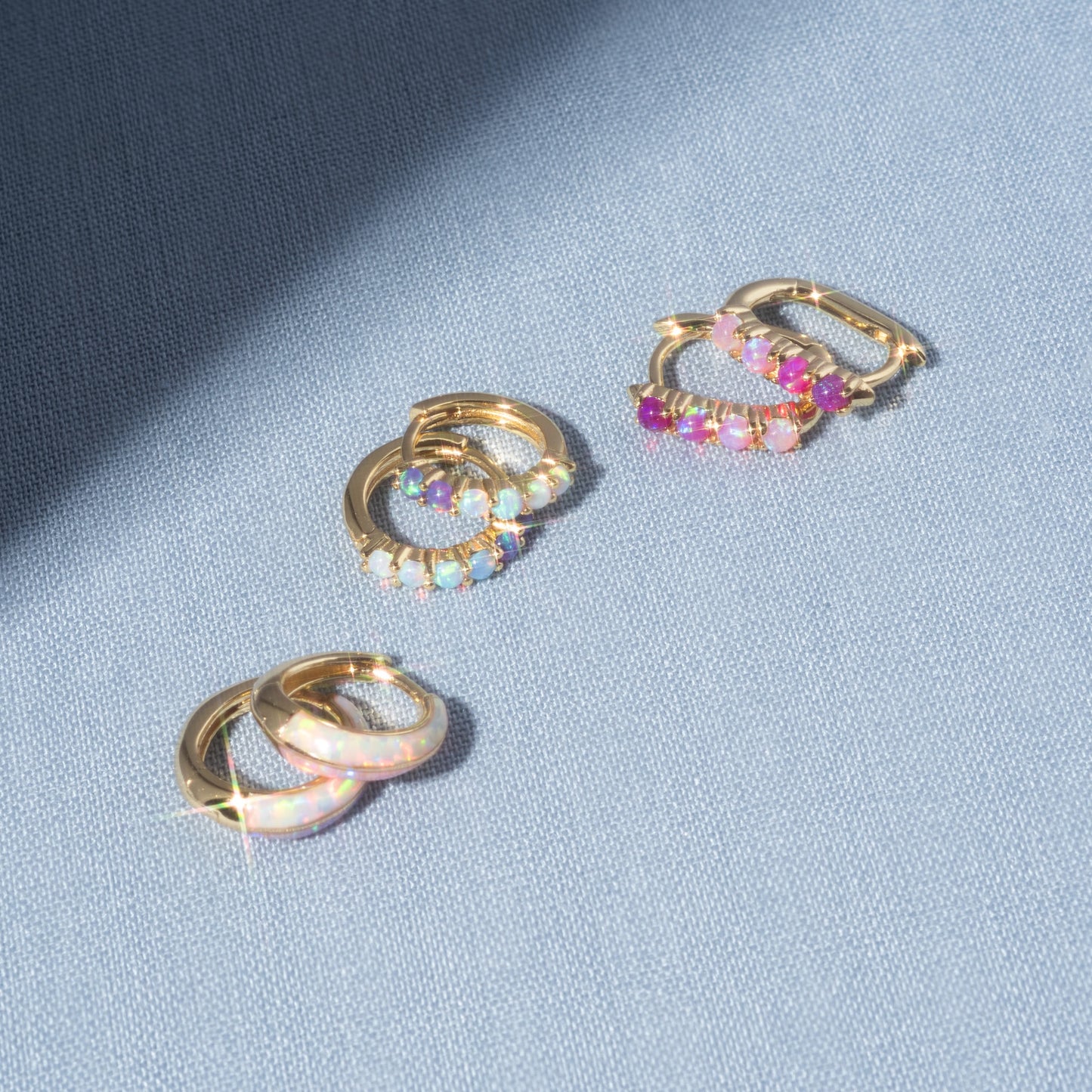 Three pairs of Opal Ombre Deux Huggies, featuring colorful gemstones and lab-created opal, rest on light blue fabric as sunlight casts gentle shadows across the scene.