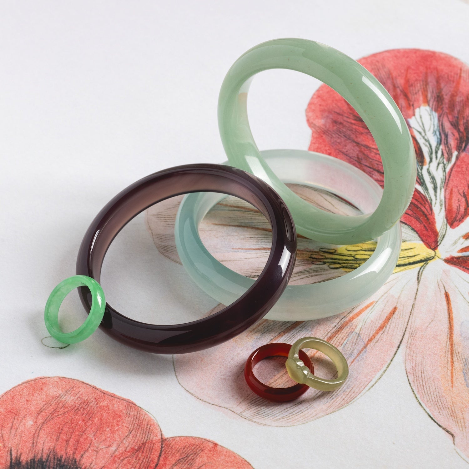 Three Heart Jade Rings in green, purple, and gold, each with a 3.5 mm band width, are arranged on a surface decorated with bold red and yellow floral artwork.