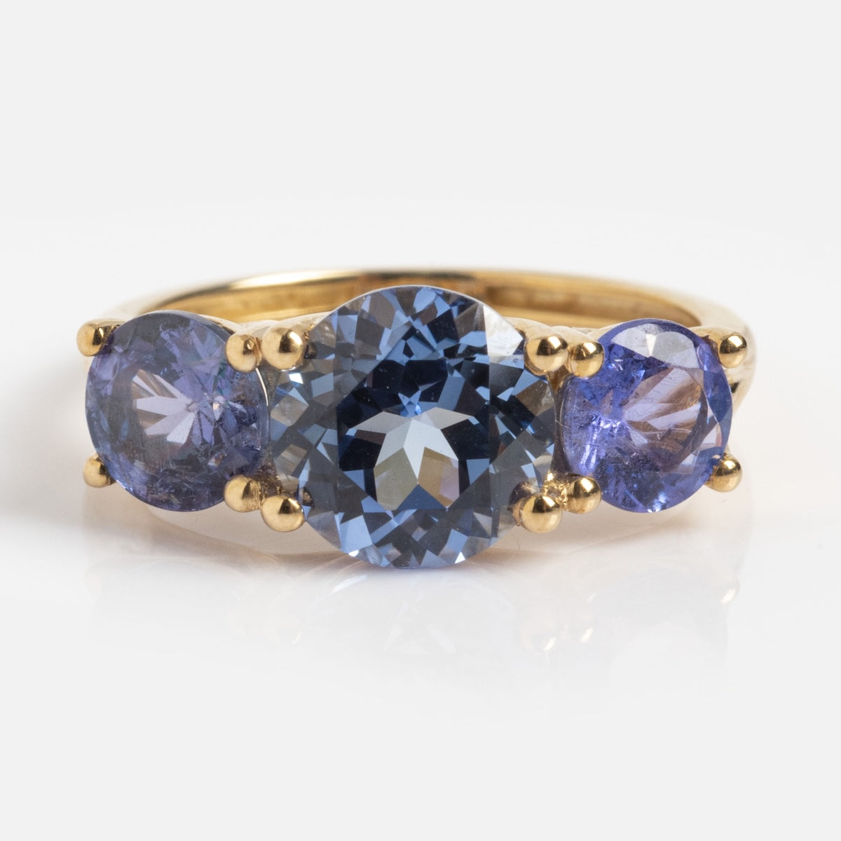The Solid Gold Trilogy Birthstone Pinky Ring shows a 7mm lab tanzanite center stone with two smaller blue gems, all set in 9k yellow gold, sample size 5, shown against a white background.