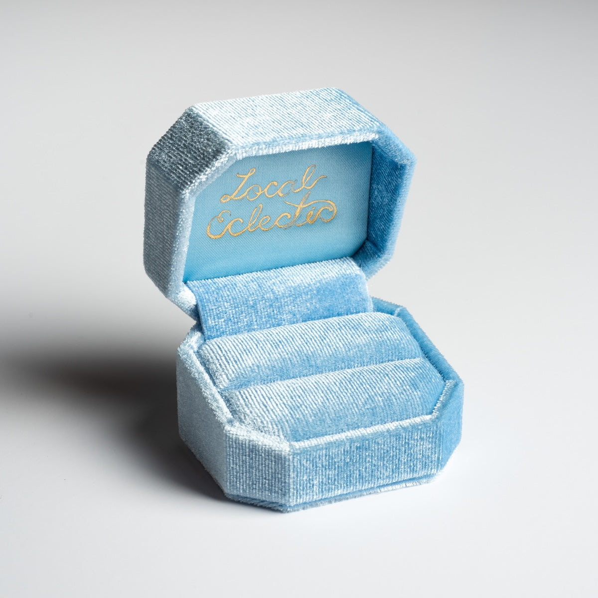 The Fancy Velvet Ring Box is a light blue, octagonal velvet box with “Local Eclectic” in gold script inside the lid. Shown open and empty on a plain background, it’s a unique choice for special occasions.