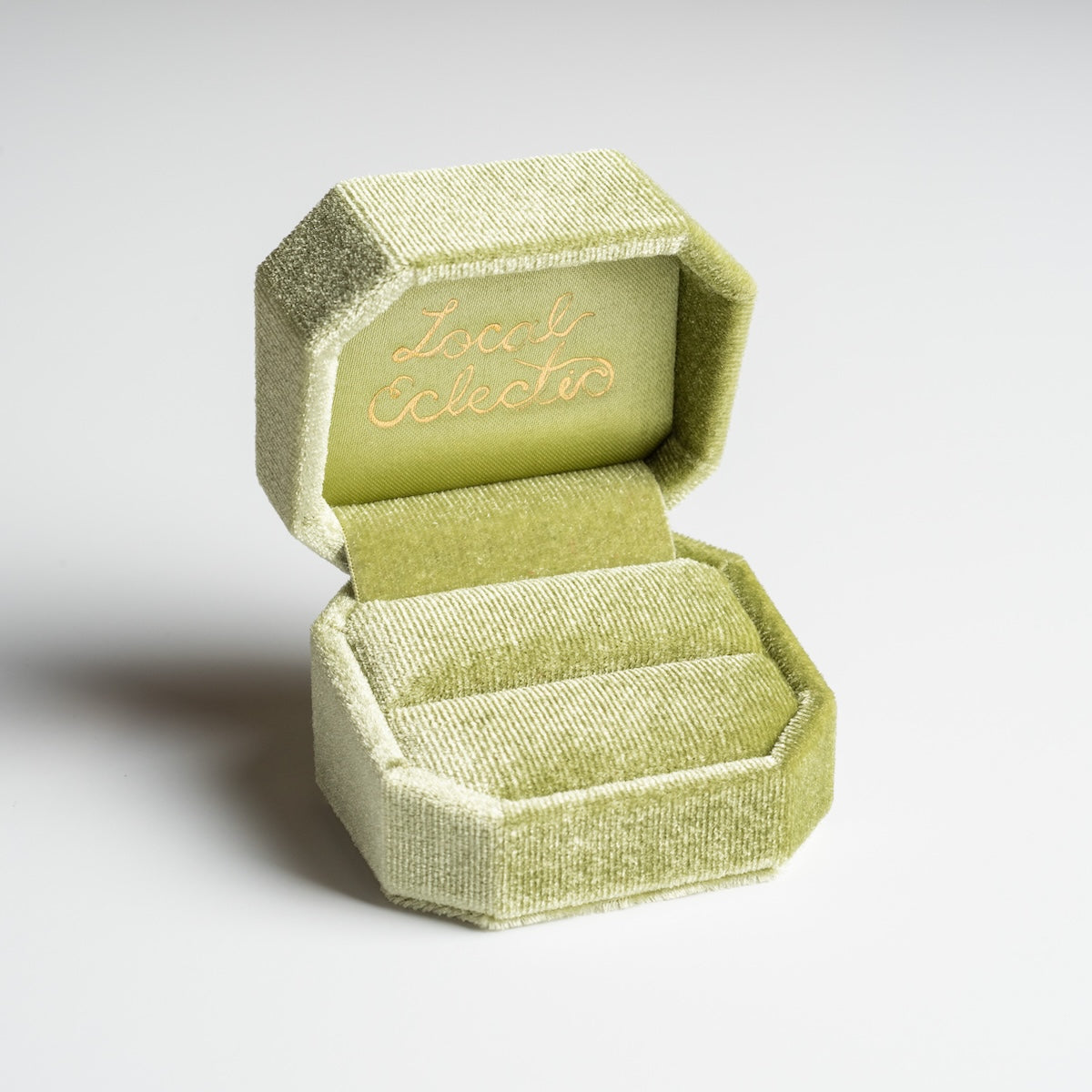 The open Fancy Velvet Ring Box features a light green velvet exterior, an octagonal shape, and gold cursive Local Eclectic text inside the lid. The empty box sits on a plain white surface.