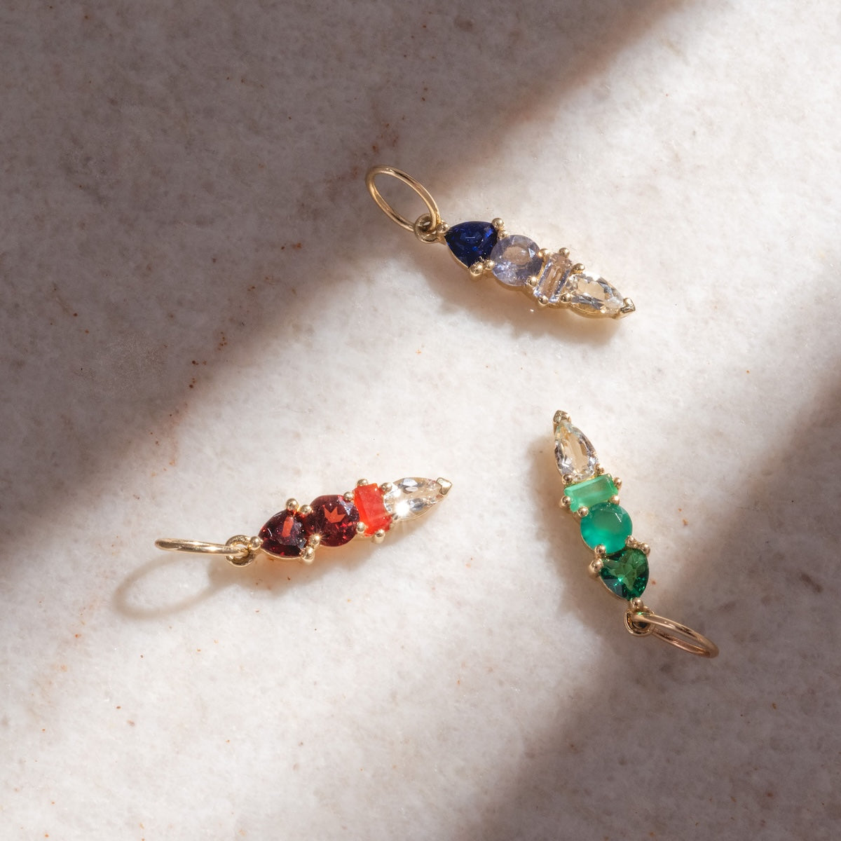 Three gold pendants, including the Solid Gold Modern Ombre Birthstone Charm set with blue, red, green, and clear gemstones, are displayed on a light textured surface under natural sunlight.