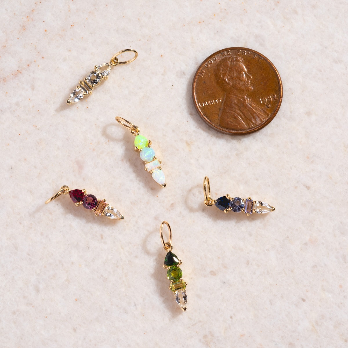 Five small, colorful Solid Gold Modern Ombre Birthstone Charms with gold loops are arranged next to a U.S. penny on a light, speckled surface to show the charm’s size for scale.