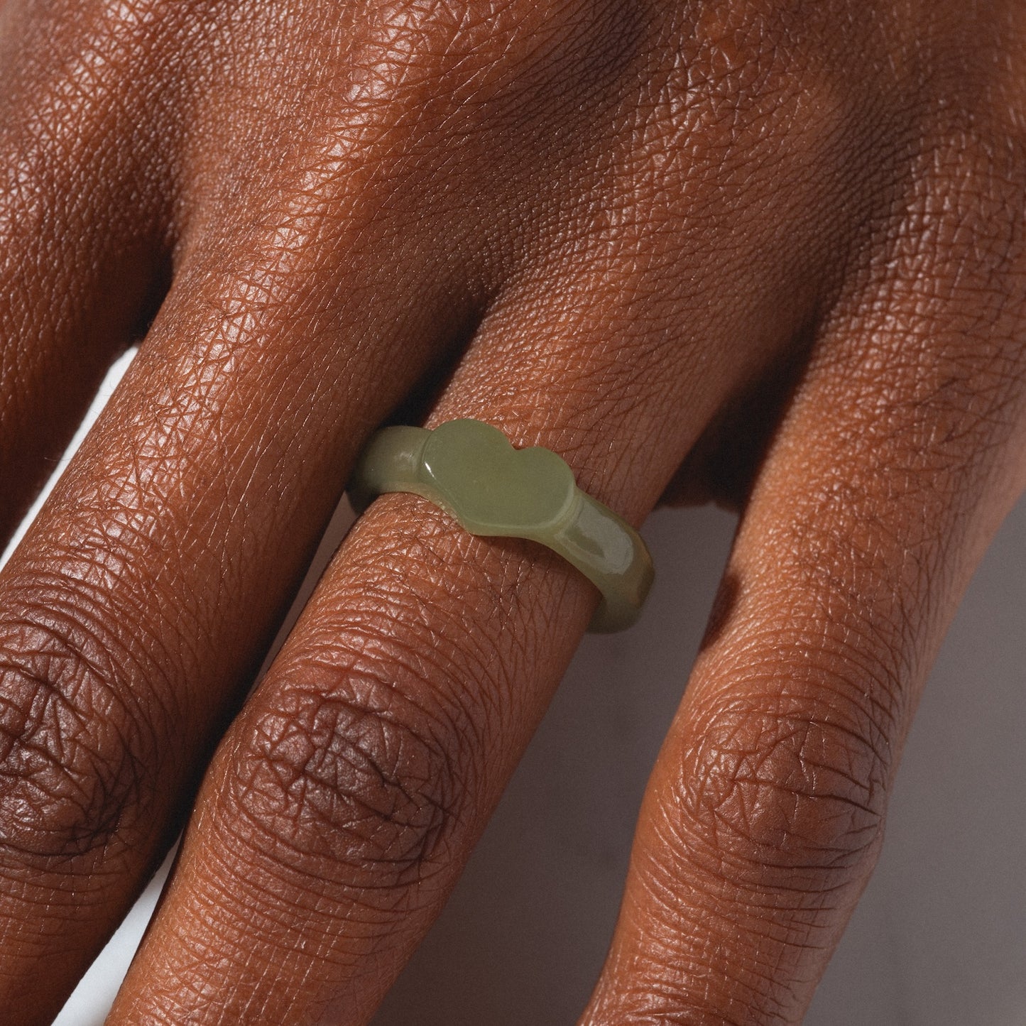 A close-up of a hand with dark skin wearing the Heart Jade Ring, a shiny, olive-green ring featuring a chunky, wavy design and a 3.5 mm band on the middle finger. The background is light and blurred.