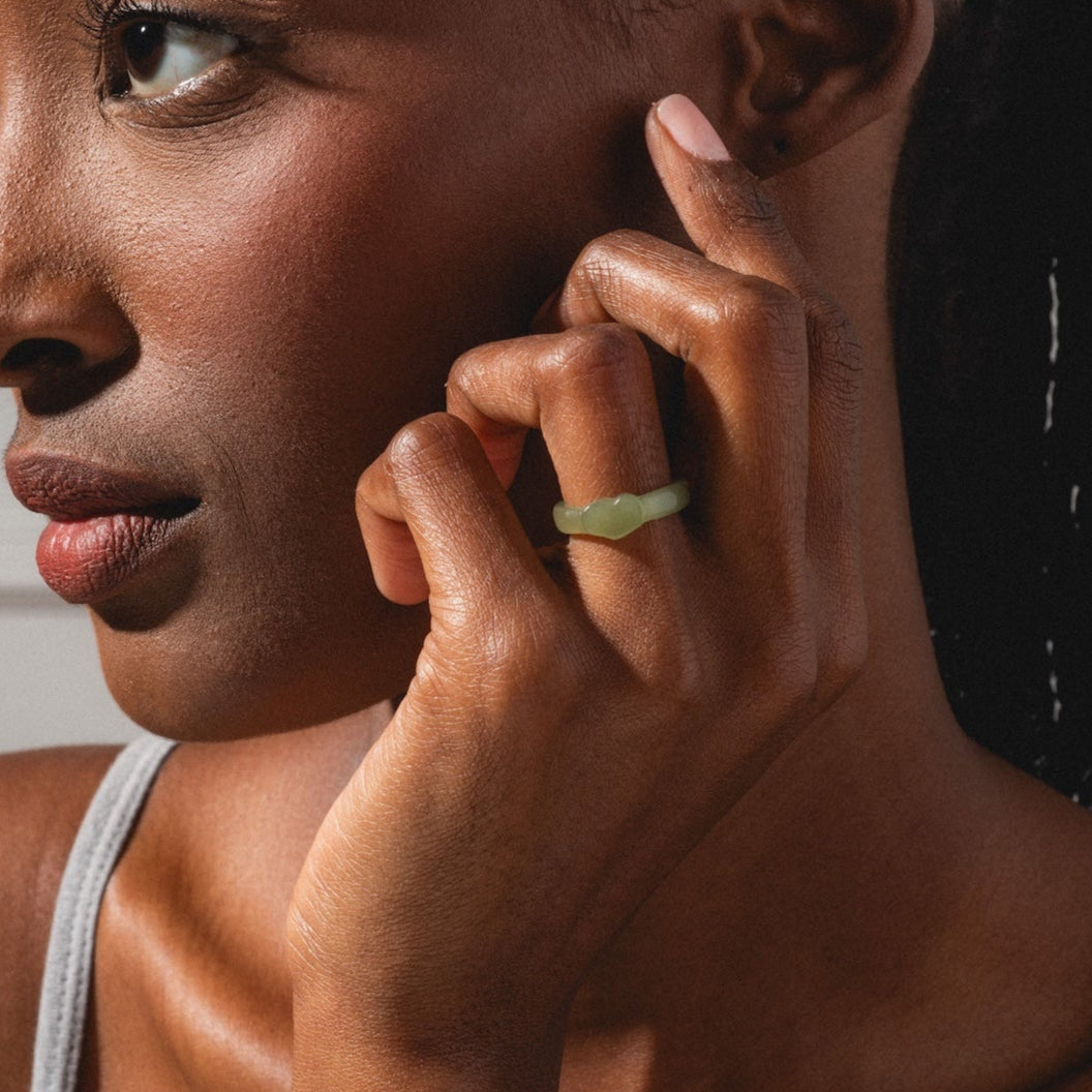 A woman with braided hair gazes to the side, her hand near her face showcasing the Heart Jade Ring with a 3.5 mm band. She wears a light-colored tank top and is softly lit.