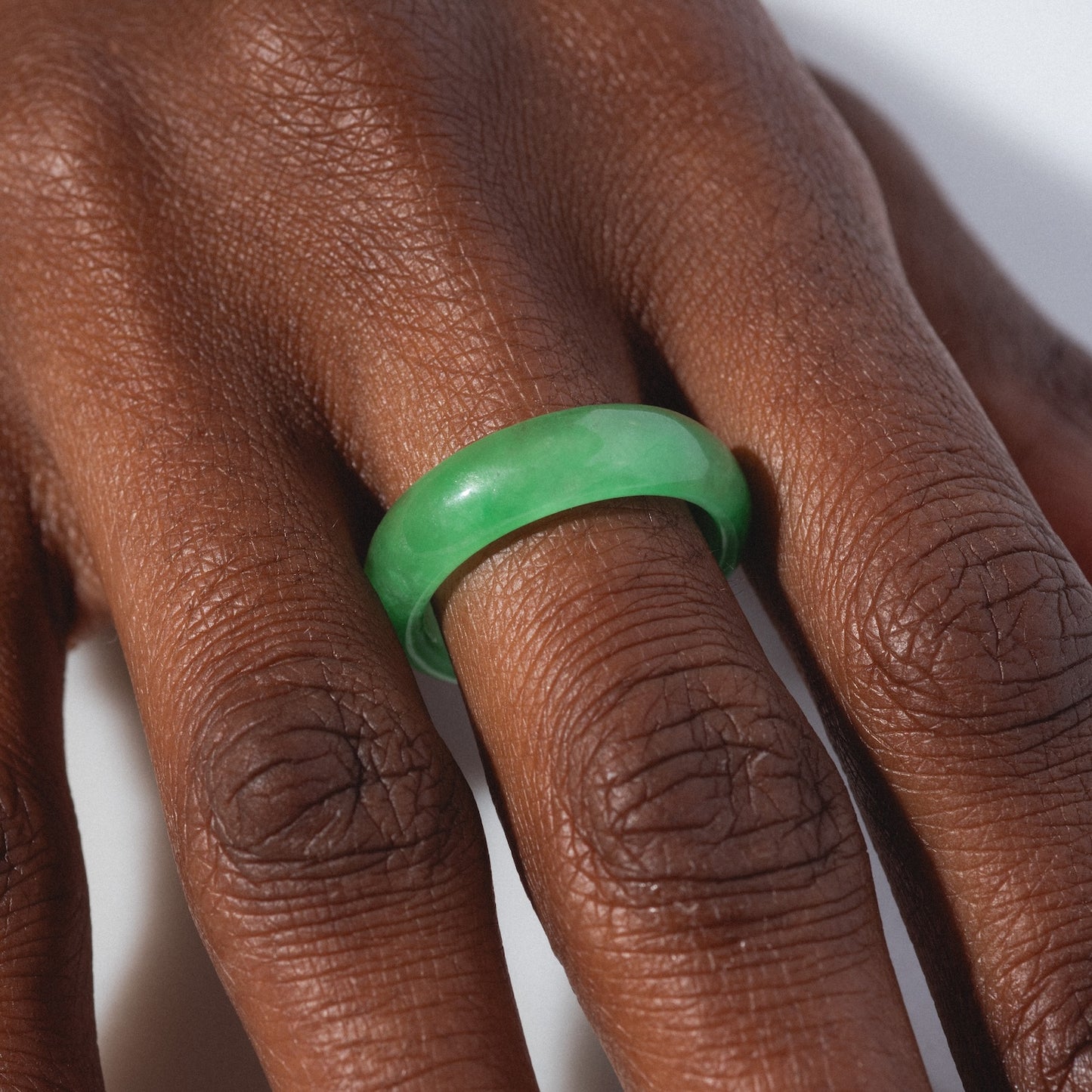 A close-up shows a hand wearing the ZZ Imperial Green Jade Ring, featuring a smooth, polished green finish and a 5 mm band on the middle finger, with natural light highlighting the ring’s shine and the skin’s texture.
