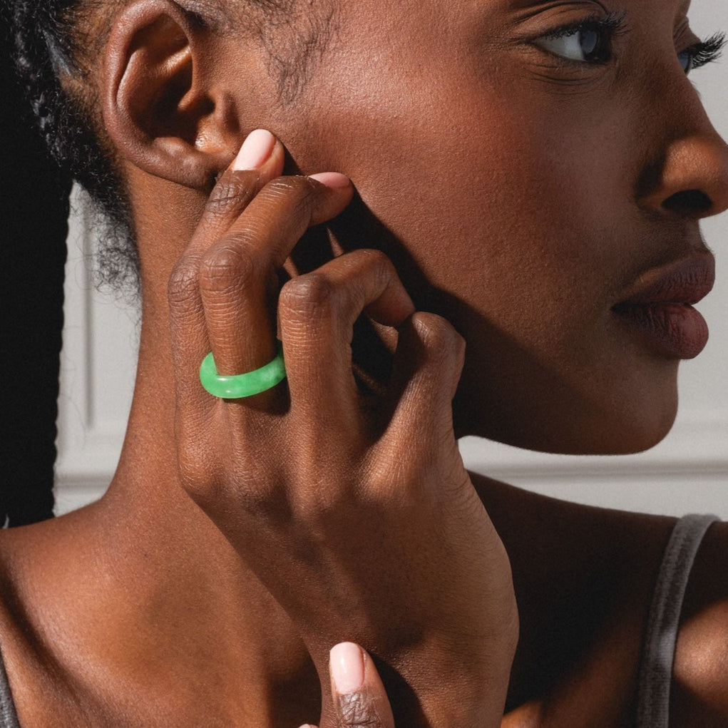 A woman with braided hair gazes to the side, her hand resting on her face. She wears a gray tank top and the ZZ Imperial Green Jade Ring with a 5 mm band on her index finger, the lighting highlighting her smooth, dark skin.