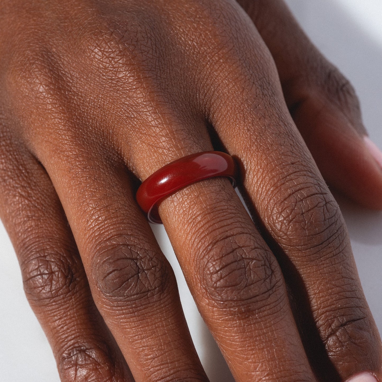 A hand wears the Persimmon Dark Orange Agate Stone Ring on the middle finger, with its glossy finish visible as the hand rests on a white surface.