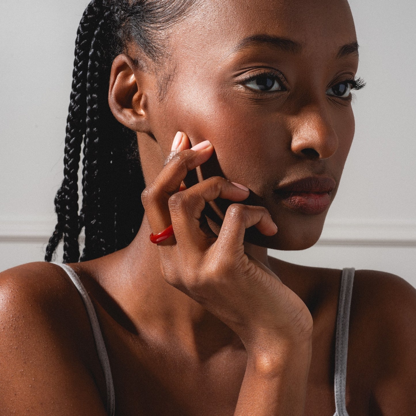 A woman with braided hair, wearing a gray strap top and the Persimmon Dark Orange Agate Stone Ring, rests her hand on her cheek and gazes thoughtfully to the side against a plain background.