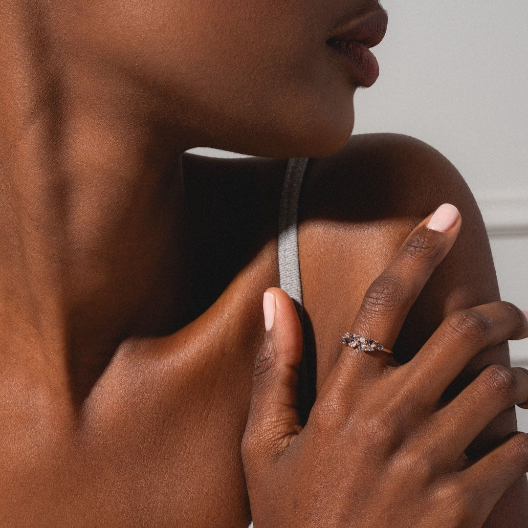 A woman with braided hair and manicured light pink nails, wearing a gray tank top, touches her collarbone to display the Gemstone Orchard Ring in gold vermeil. She is turned slightly to the side against a neutral background.
