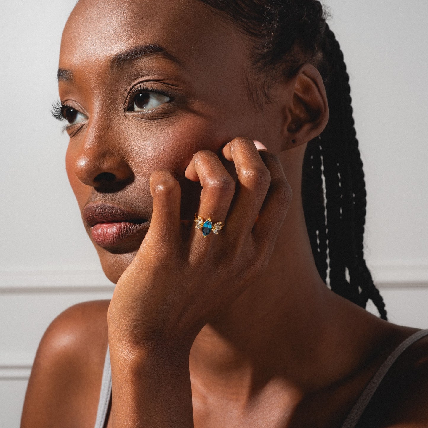 A woman with braided hair rests her chin on her hand, wearing the Gemstone Firefly Ring in yellow gold vermeil with a London blue topaz. She gazes thoughtfully to the side, softly lit against a plain background.