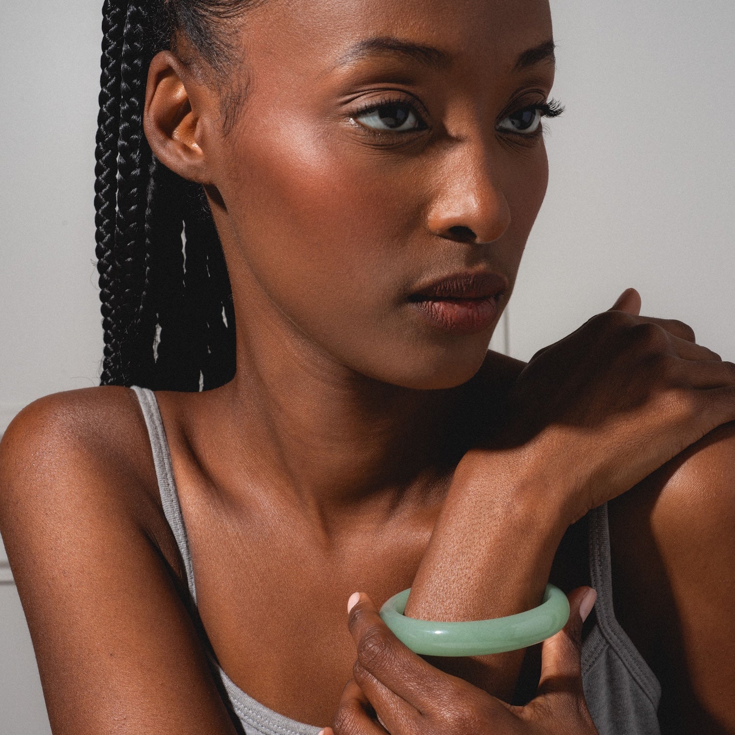 A woman with braided hair, wearing a grey tank top and a Smoke Green Jade Quartzite Stone Bangle, gazes thoughtfully to the side with her hand on her shoulder against a neutral background.