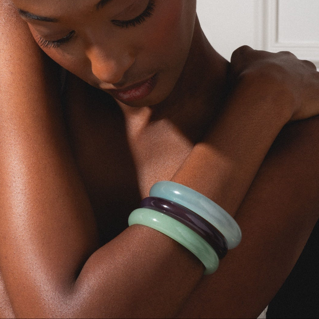 A woman with braided hair hugs her shoulders, looking down, wearing a wide silver cuff and the Moon Purple Jade Stone Bangle in dark purple on her right wrist, set against a neutral background.